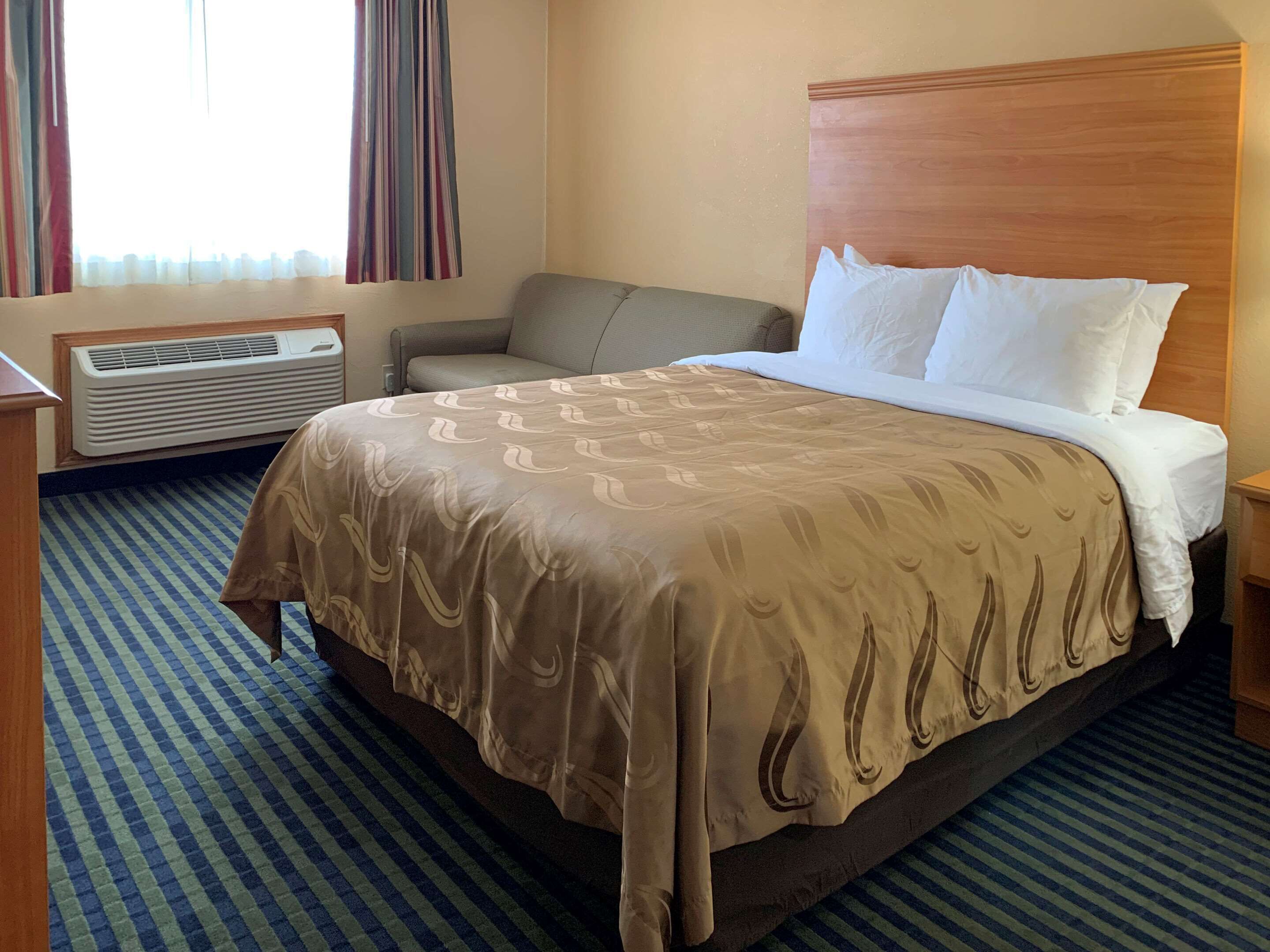 A hotel room with a large bed, a bedside table, a chair, a window with curtains, and a wall-mounted air conditioner.