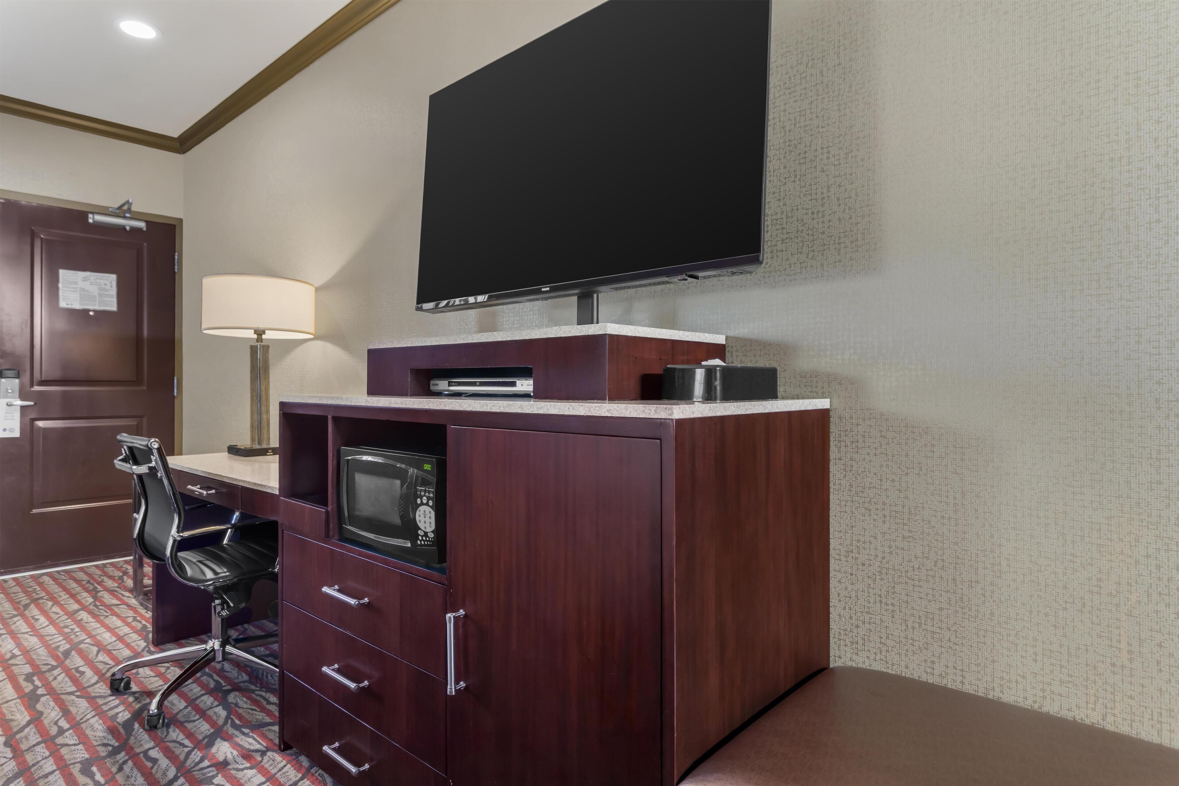 A hotel room with a wooden desk, a flat-screen TV mounted on the wall, a desk lamp, a chair, and a patterned carpet.