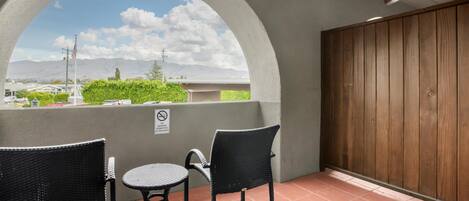 Un balcón al aire libre con dos sillas de mimbre, una mesa redonda pequeña, un muro de madera y vistas a una ciudad y montañas.