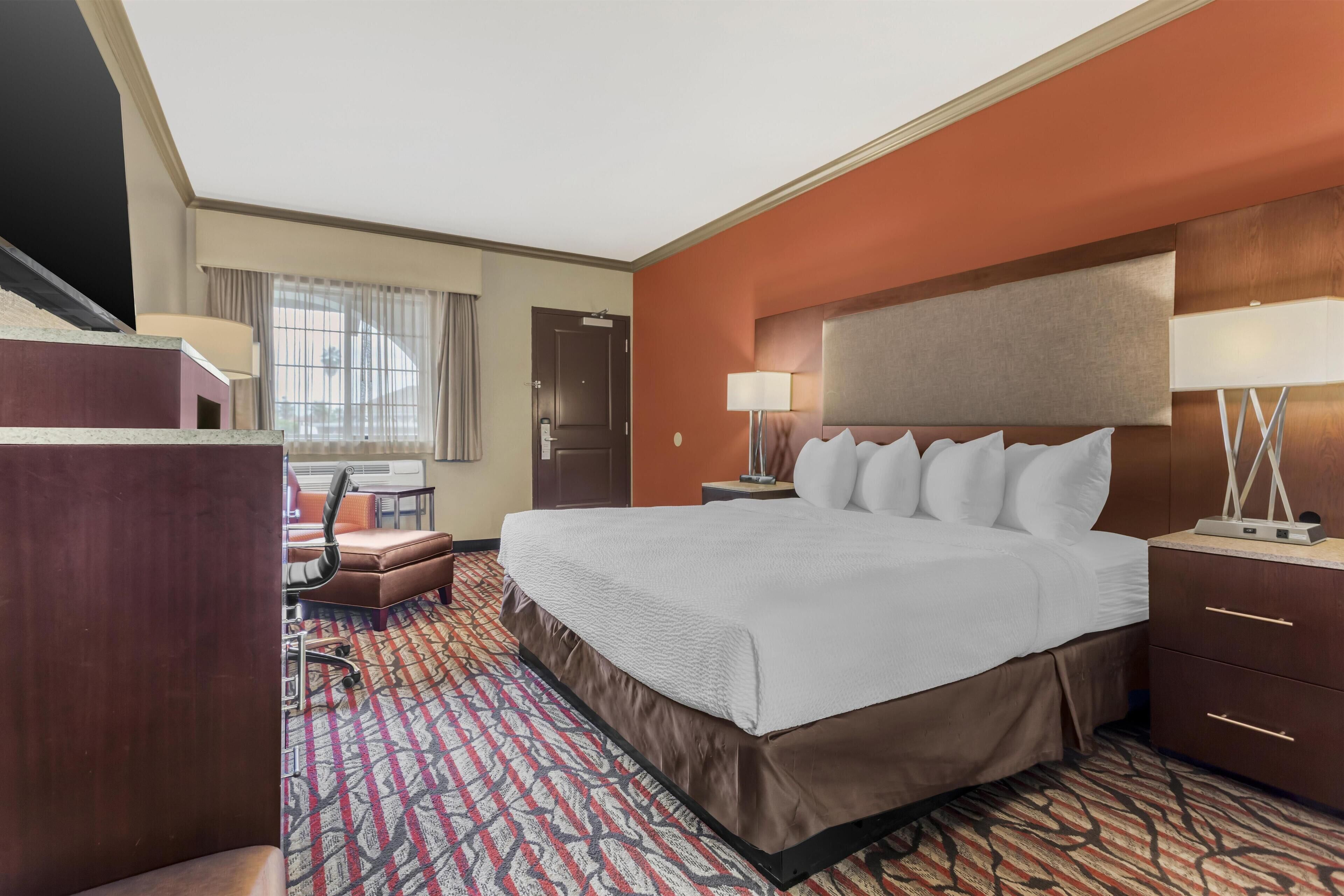 A hotel room with a large bed, a desk with a chair, a lamp, and a window with curtains.