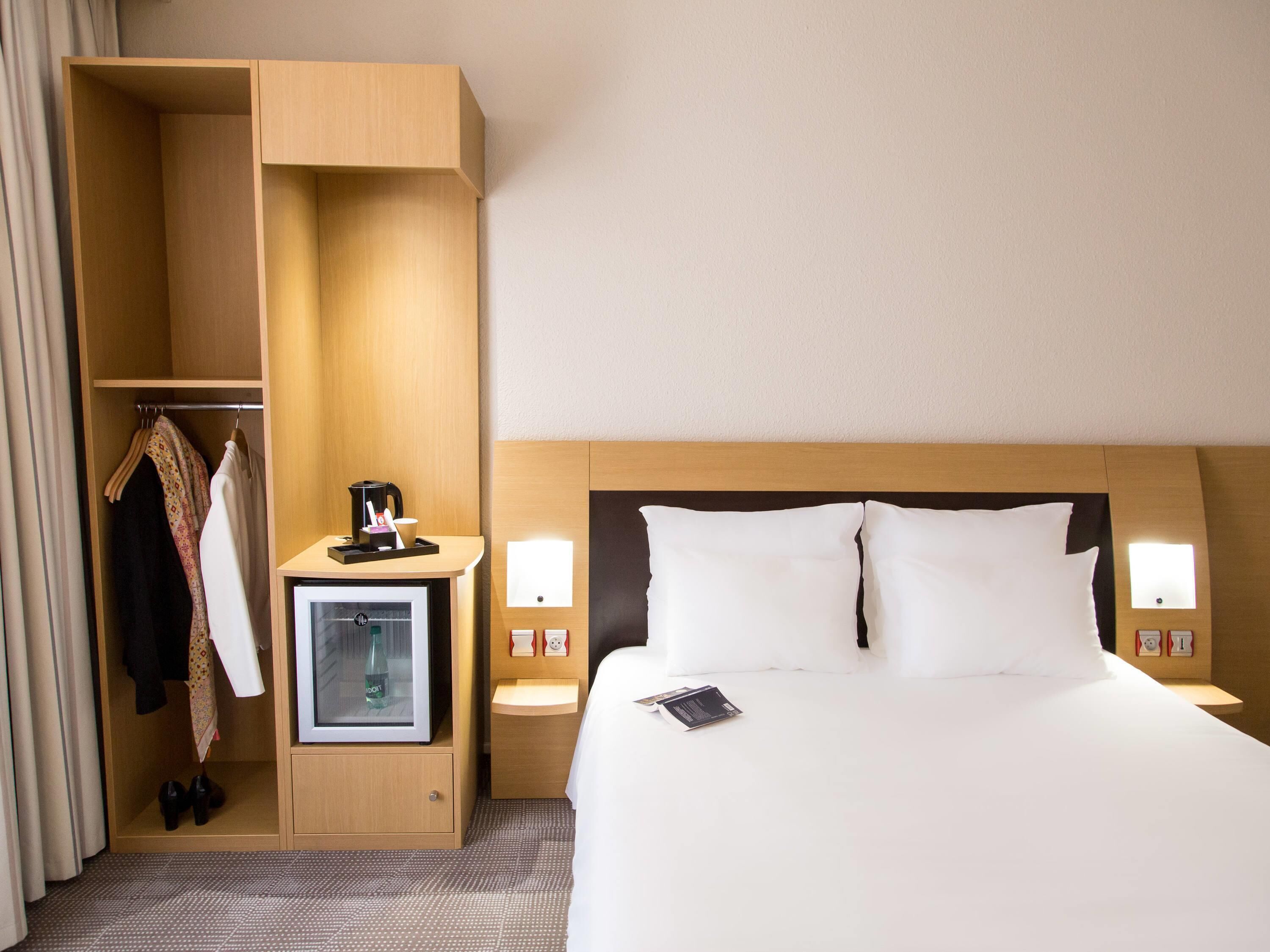 Ein Hotelzimmer mit Bett, Kleiderschrank, Minibar und einem kleinen Schreibtisch mit Lampe.