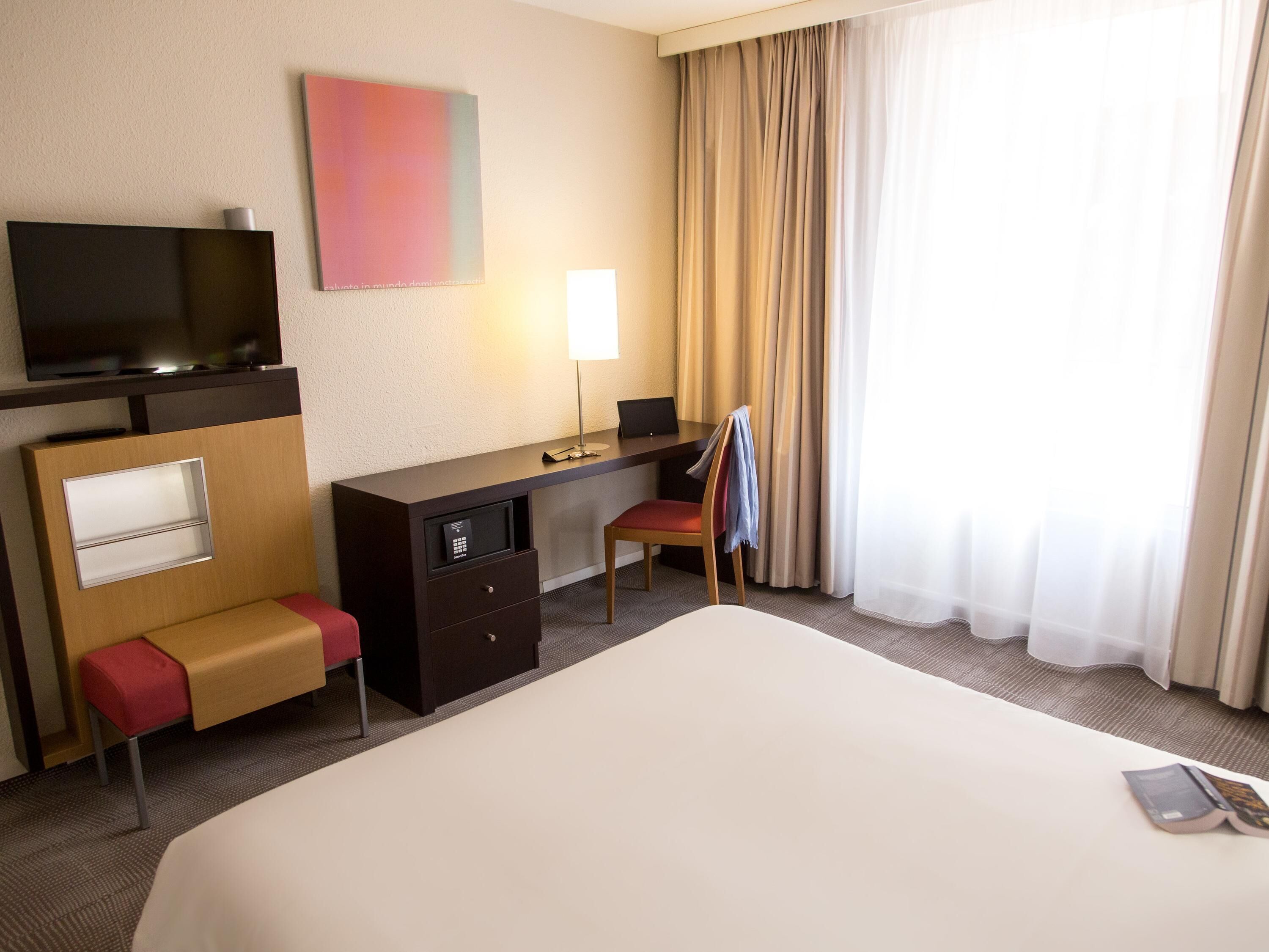 A hotel room with a bed, a desk, a chair, a TV, and a window with curtains.