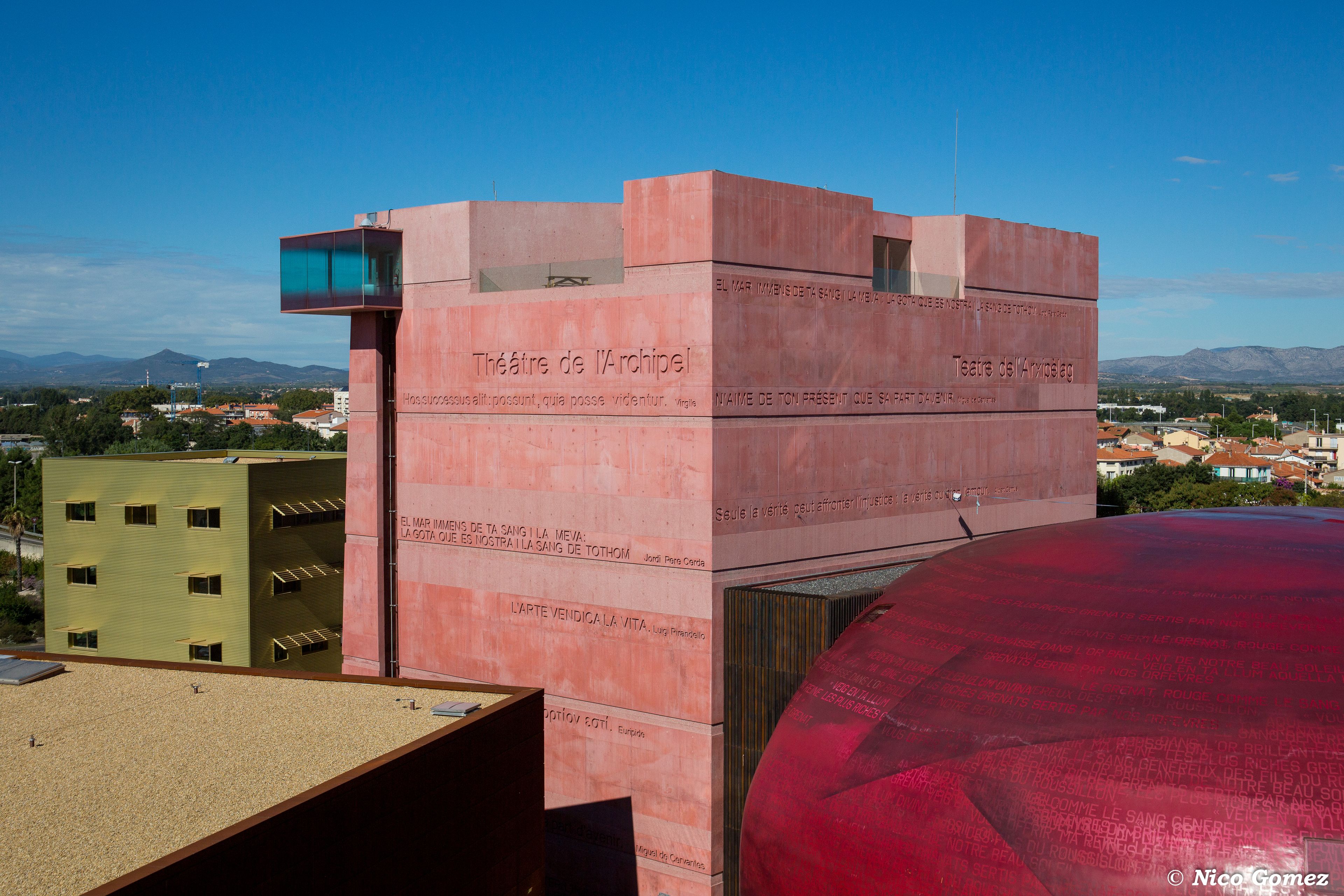 Un bâtiment moderne dont la façade porte l’inscription « Théâtre de l’Archipel », caractérisé par une architecture originale avec une partie saillante et une grande structure en forme de dôme rouge située à proximité.