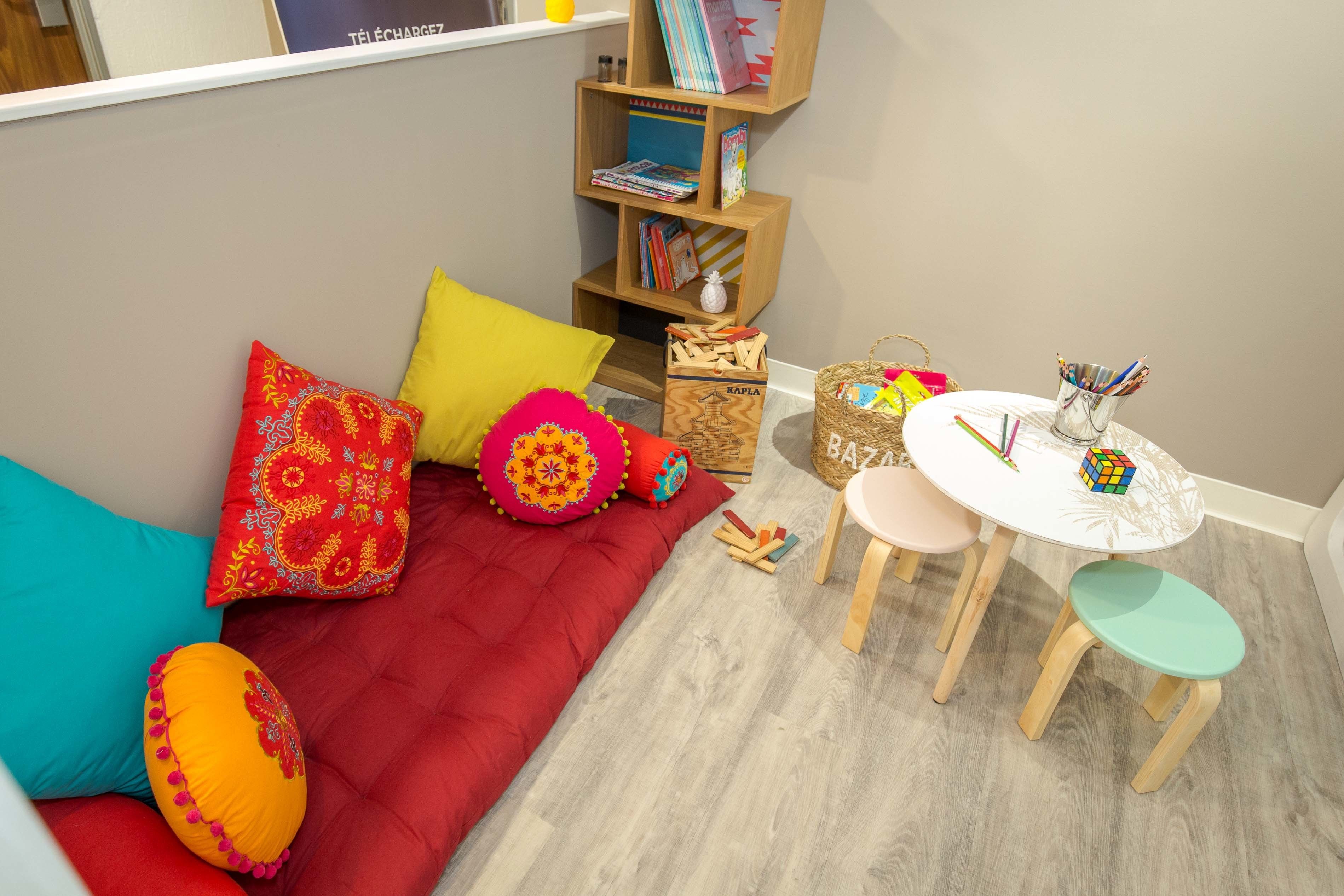 Una habitación acogedora con un sofá rojo, almohadones coloridos, una pequeña mesa de madera con sillas y una estantería llena de libros y objetos decorativos.