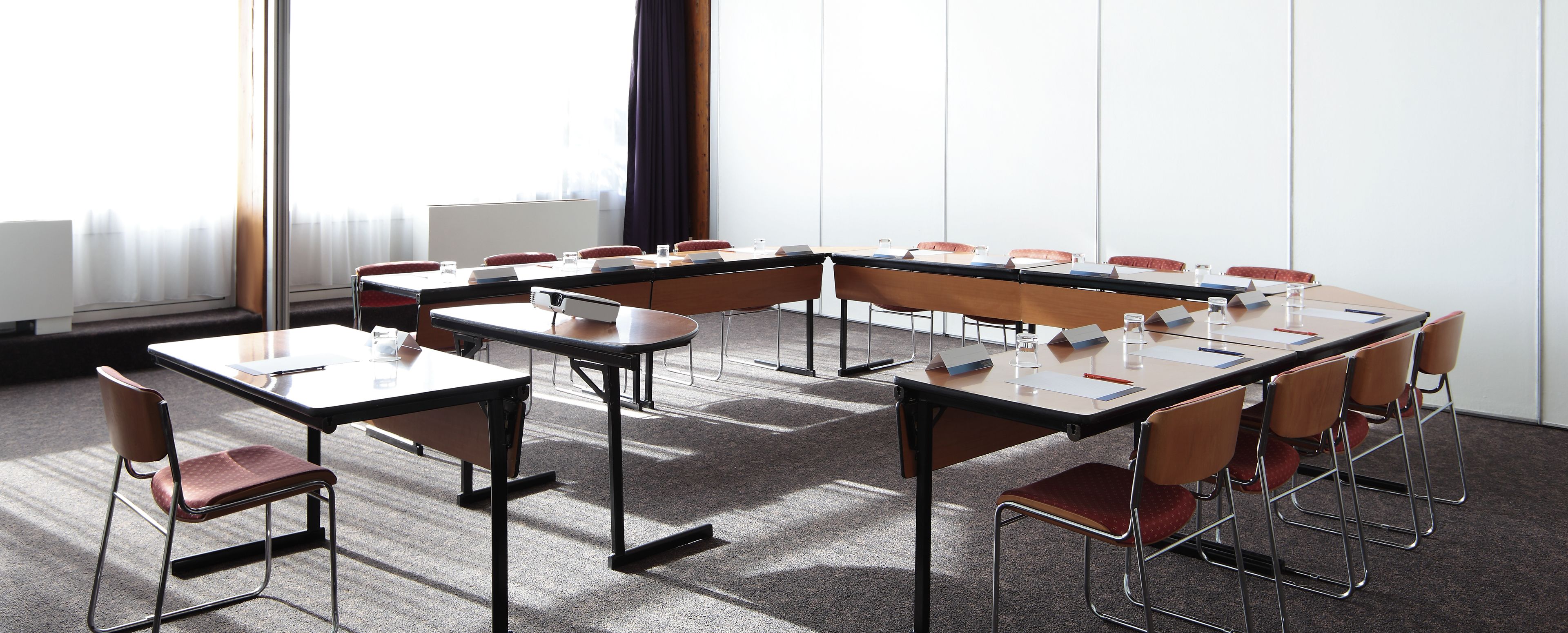 Une salle de conférence dotée de plusieurs tables et de chaises, de grandes fenêtres avec des rideaux et d’un sol recouvert de moquette.