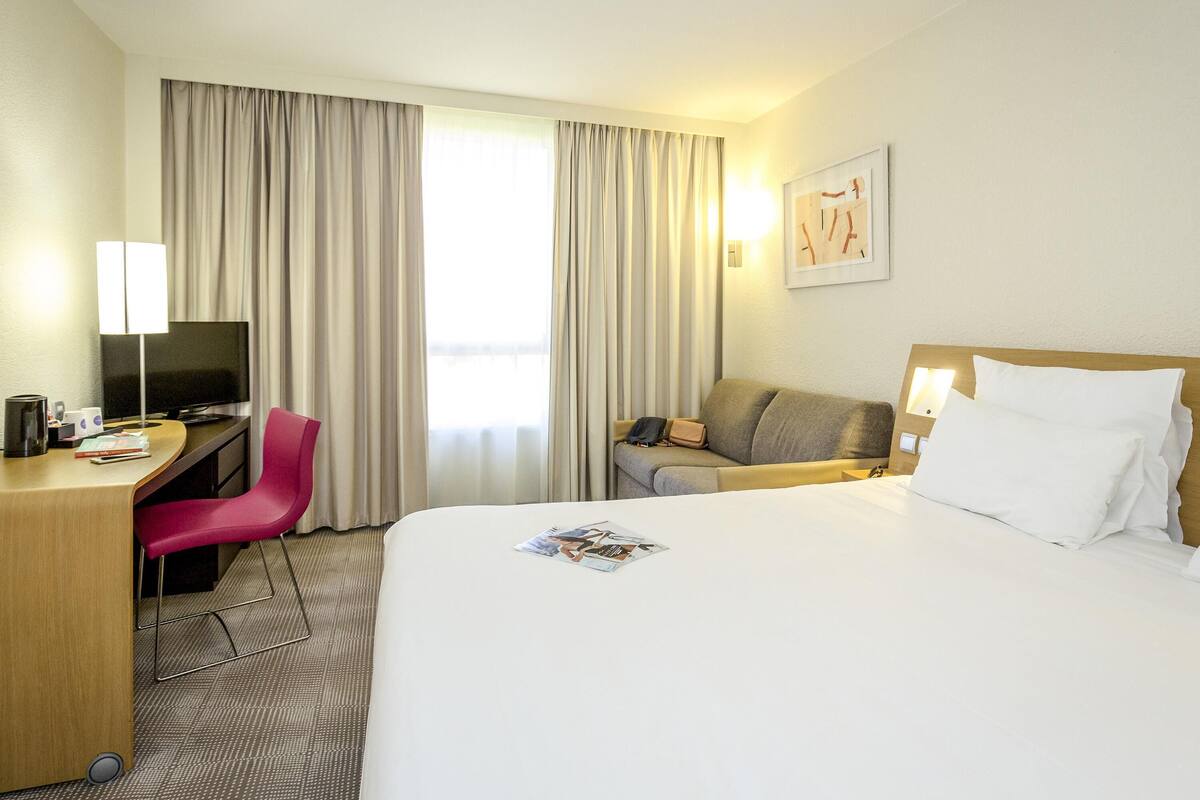A hotel room with a bed, a desk with a TV, a chair, a sofa, and a window with curtains.