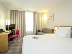 A hotel room with a bed, a desk with a TV, a chair, a sofa, and a window with curtains. - Novotel Perpignan Nord Rivesaltes (Rivesaltes)