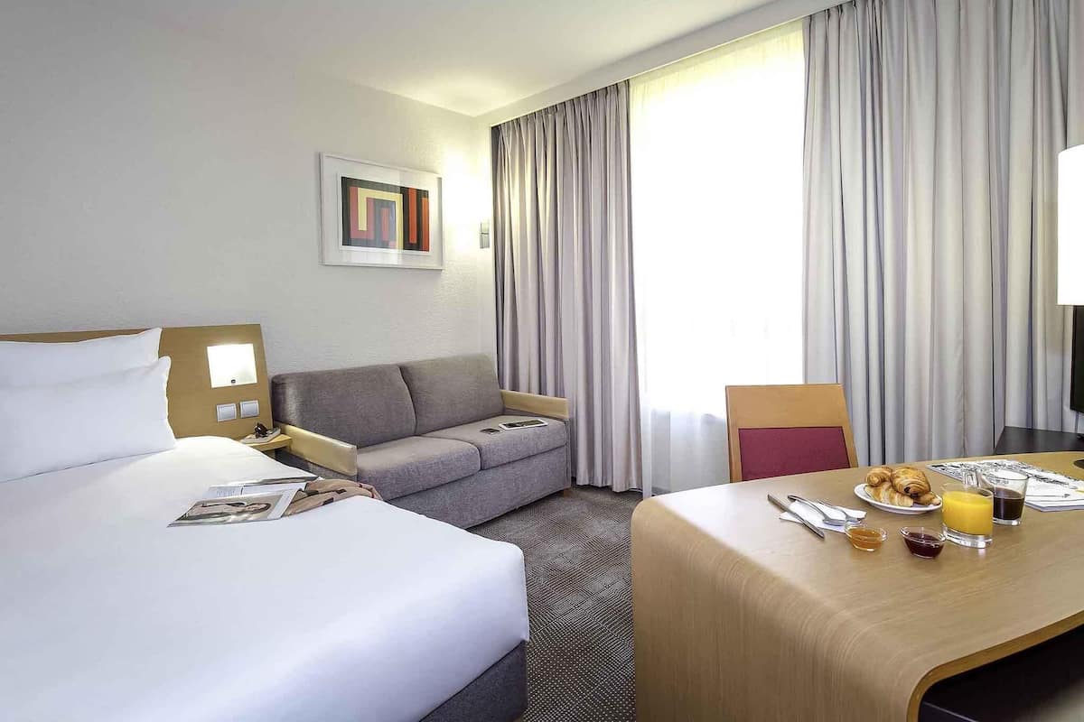 A hotel room with a bed, a sofa, a dining table with breakfast, and a large window with curtains.