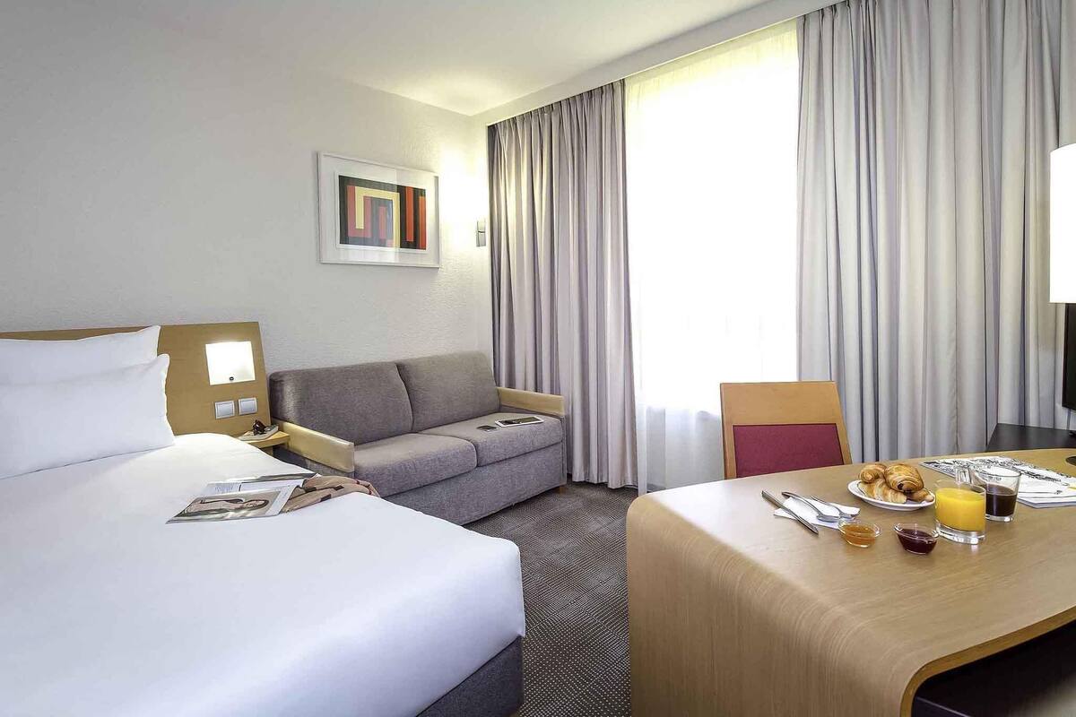 A hotel room with a bed, a sofa, a dining table with breakfast, and a large window with curtains.