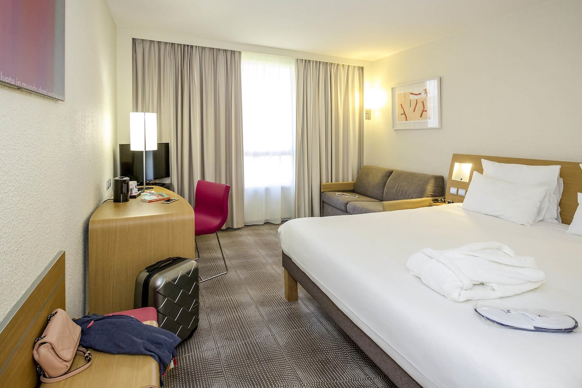 A hotel room with a bed, a desk, a chair, a sofa, and a suitcase.
