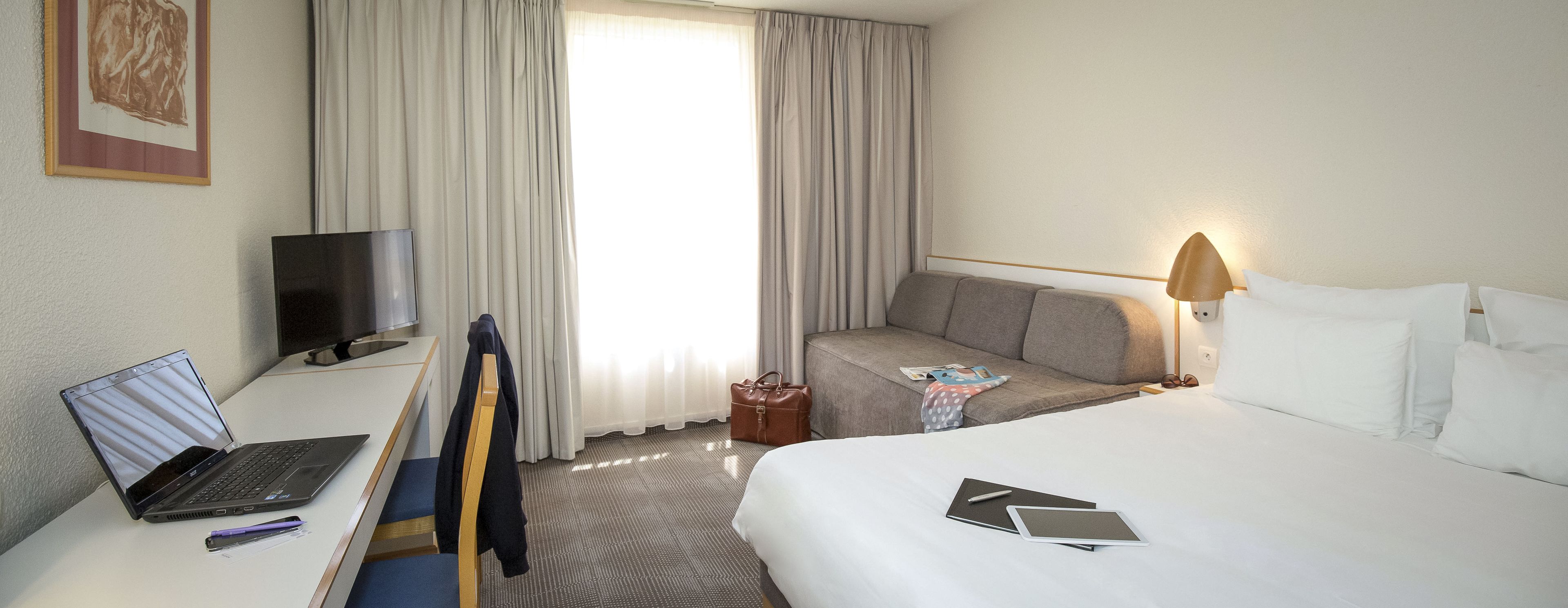A hotel room with a bed, a desk with a laptop, a chair, a sofa, a lamp, and a TV.