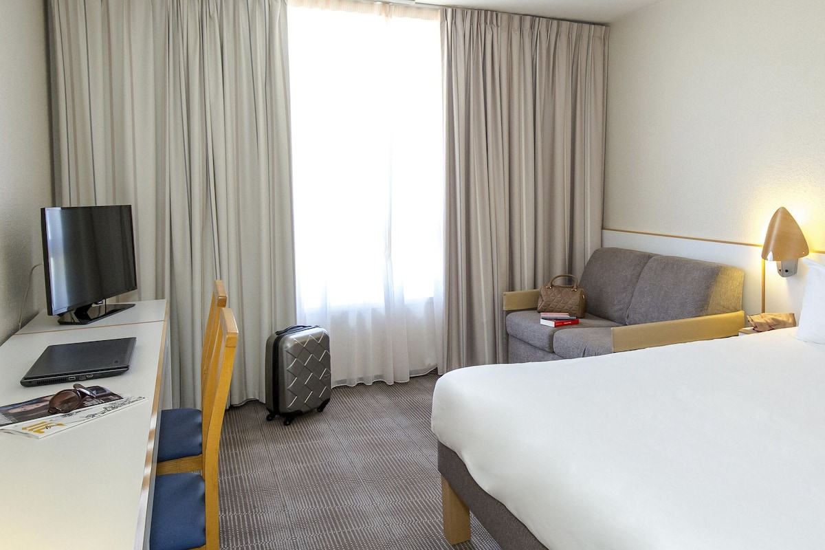 Ein Hotelzimmer mit Bett, Schreibtisch mit Laptop, Stuhl, Koffer, Fernseher und Nachttisch mit Lampe.