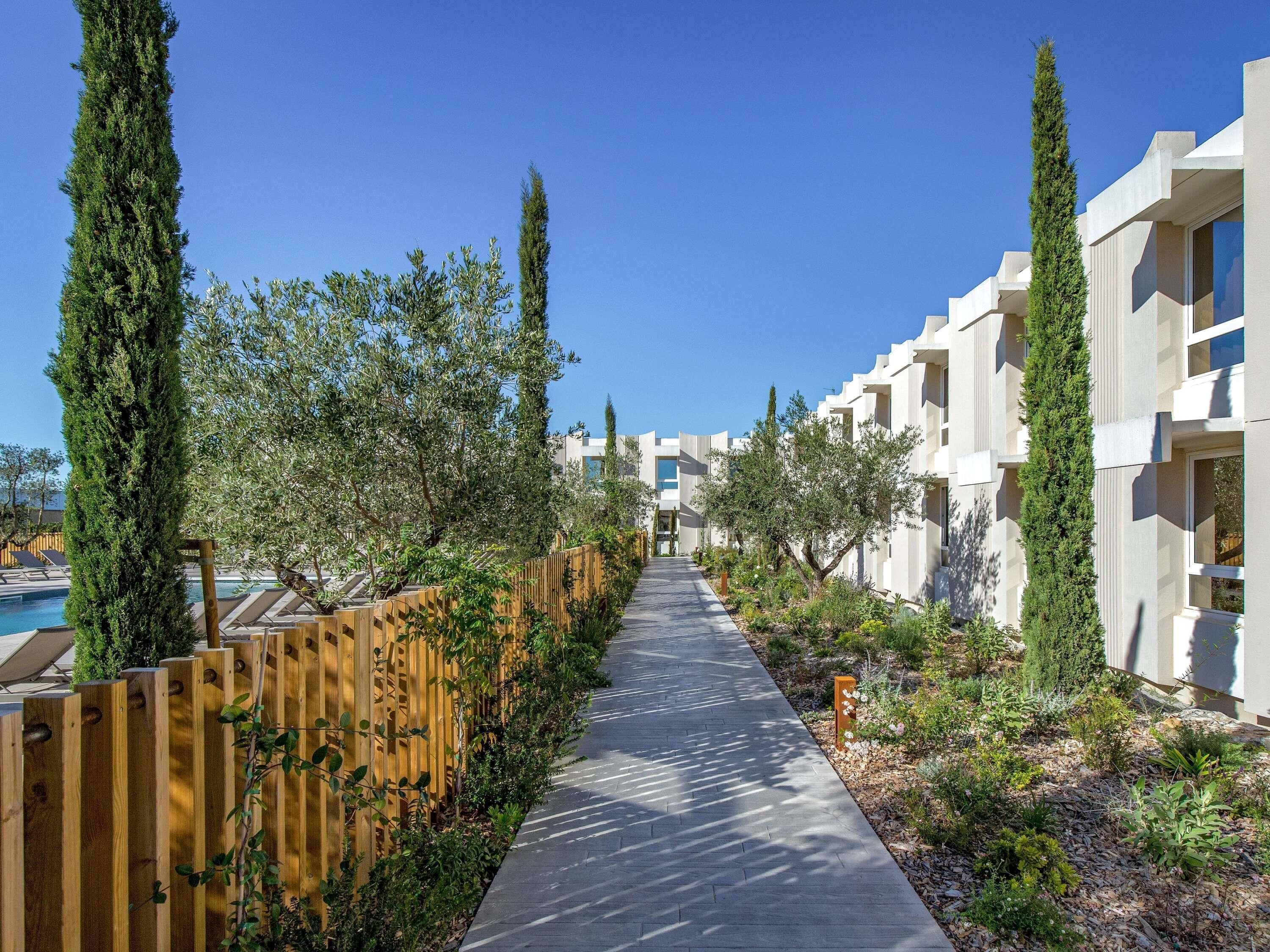 Un sendero bordeado de árboles y vallas de madera que conduce a una zona de piscina, con edificios modernos a ambos lados.