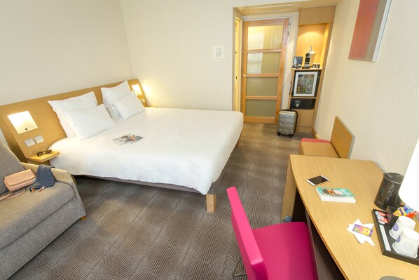 A hotel room with a bed, a desk, a chair, a small table, a suitcase, and a nightstand with a lamp.