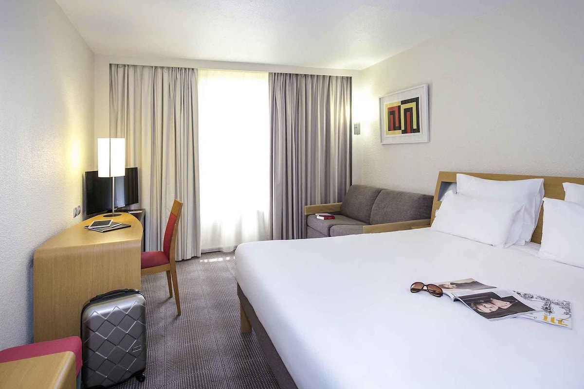Ein Hotelzimmer mit Bett, Schreibtisch, Stuhl, Fernseher und einem Fenster mit Vorhängen.