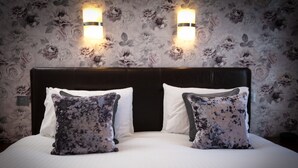 Cama bem arrumada com duas almofadas decorativas, cabeceira escura e papel de parede floral, com arandelas.