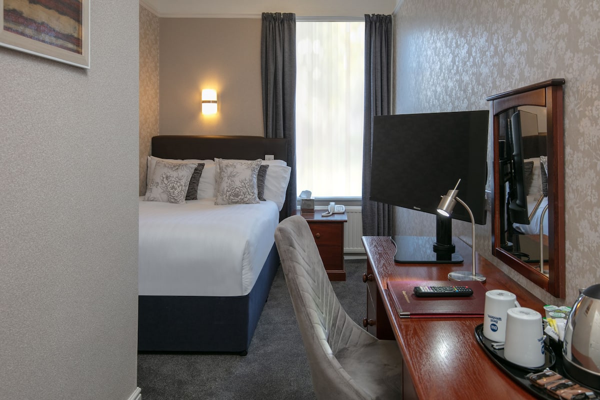 A hotel room with a bed, bedside table, lamp, mirror, and a view of the outside.