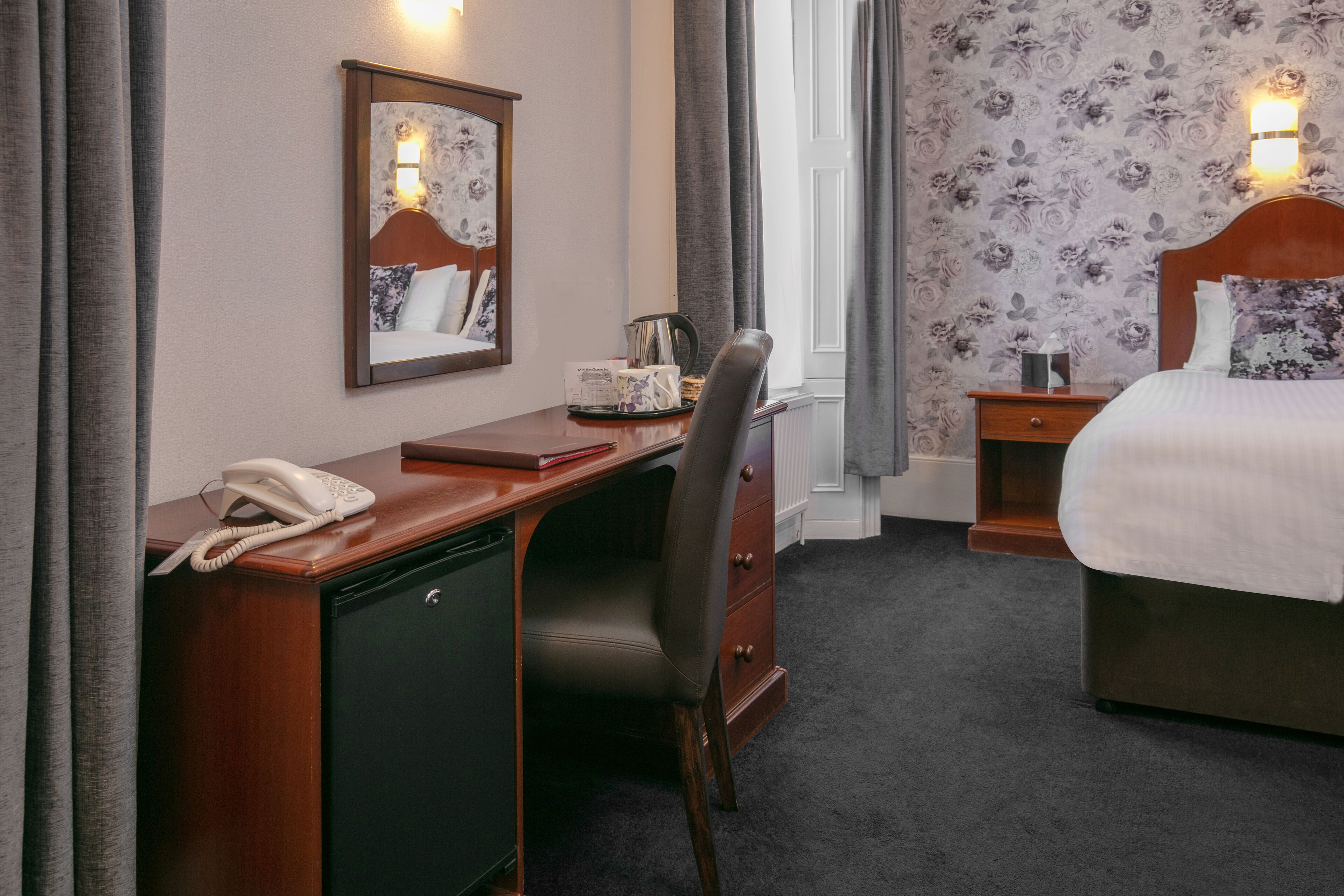 A hotel room with a wooden desk, a chair, a bed, a nightstand, a mirror, and a window with curtains.