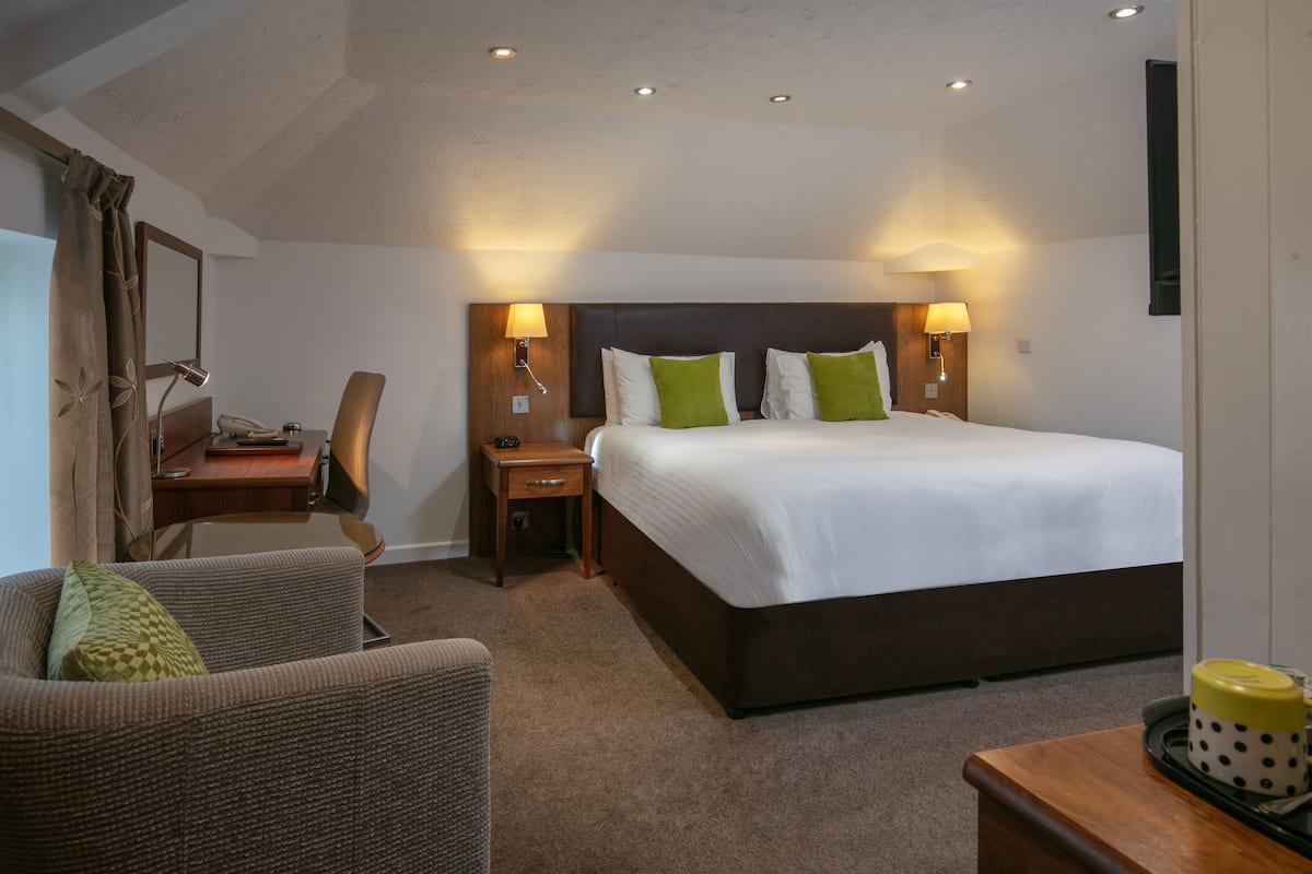 A hotel room with a large bed, two bedside lamps, a desk with a telephone, a chair, and a small table with a mug.