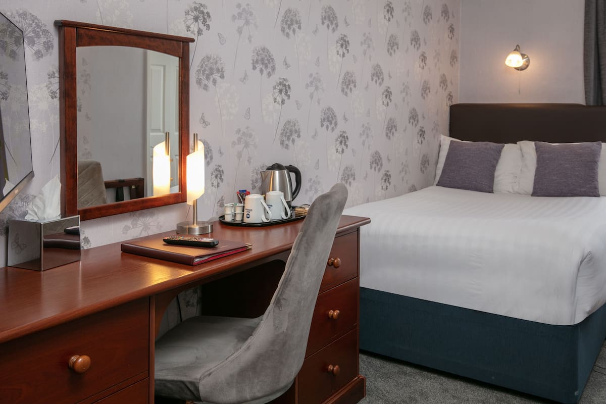 A hotel room with a wooden desk, a chair, a bed with white and blue bedding, a nightstand with a lamp, a mirror, and a floral wallpaper pattern.