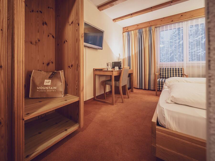 A hotel room with a wooden desk, a bed, a chair, and a window with curtains.