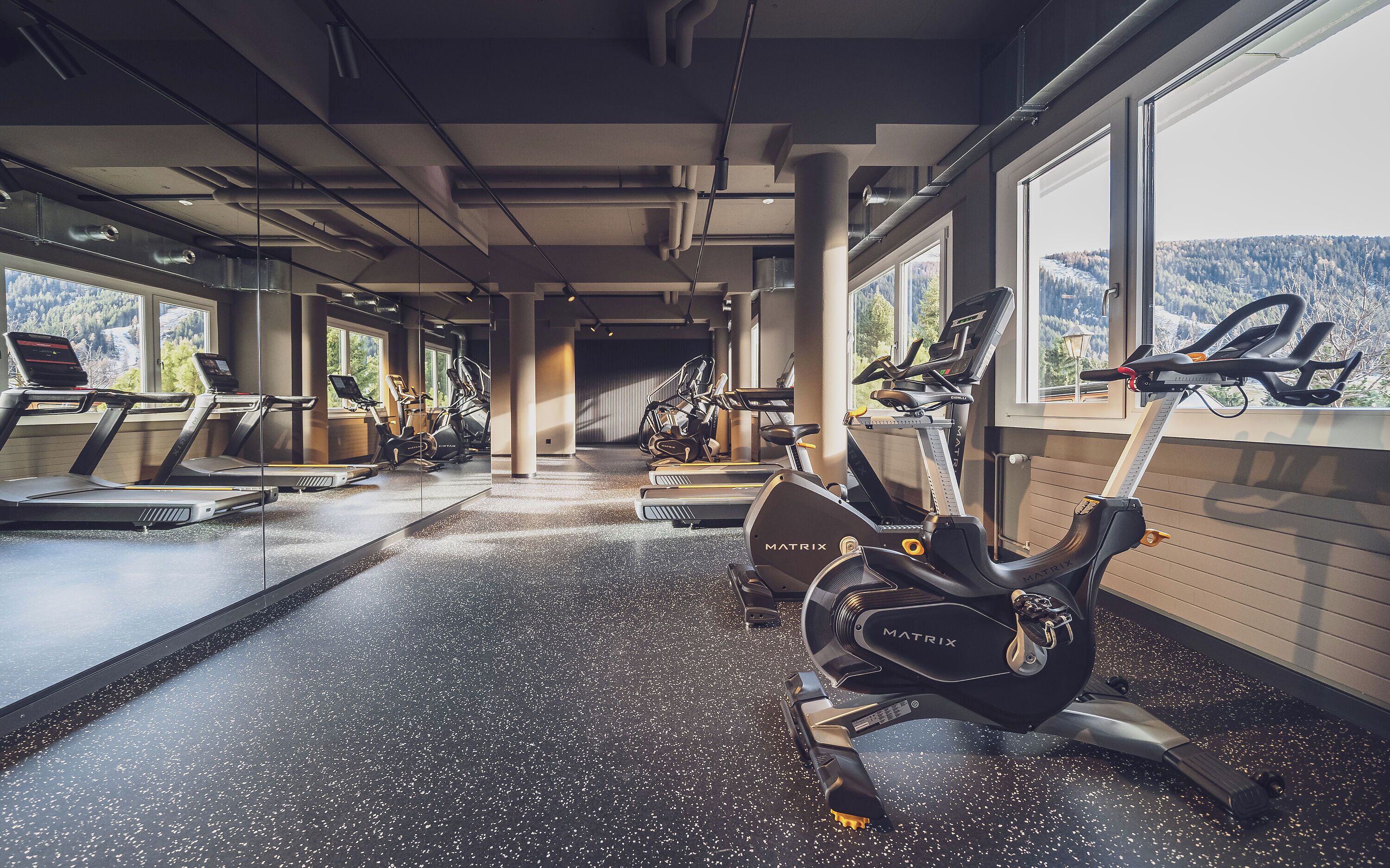 A gym with treadmills, elliptical machines, and stationary bikes, with large windows offering a view of a snowy landscape.