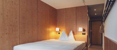 Habitación de hotel con paredes de paneles de madera, una cama con ropa blanca, dos lámparas fijadas a la pared y una escalera a la derecha.