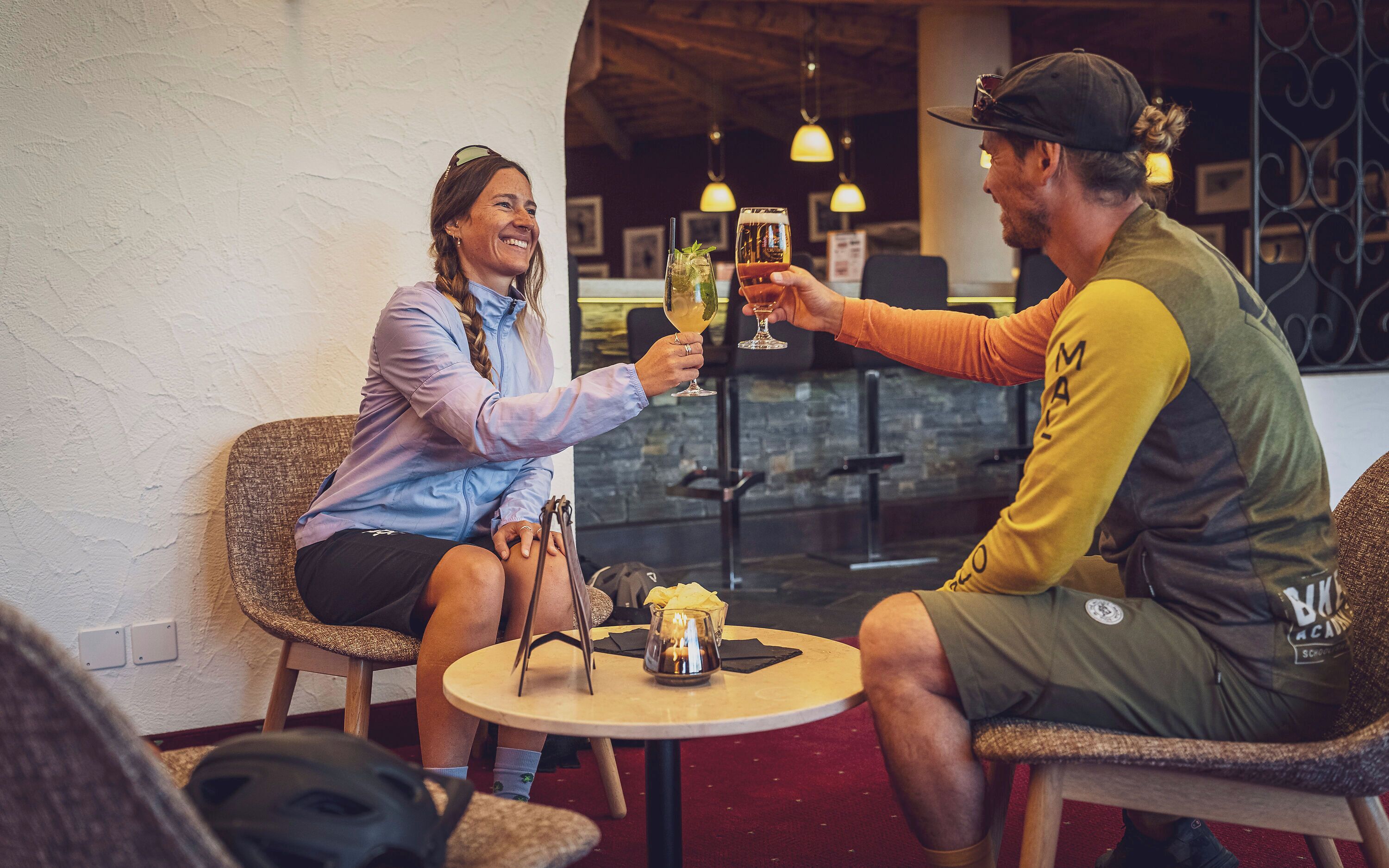 Two people toasting with drinks at a table in a restaurant with a brick wall and hanging lights.