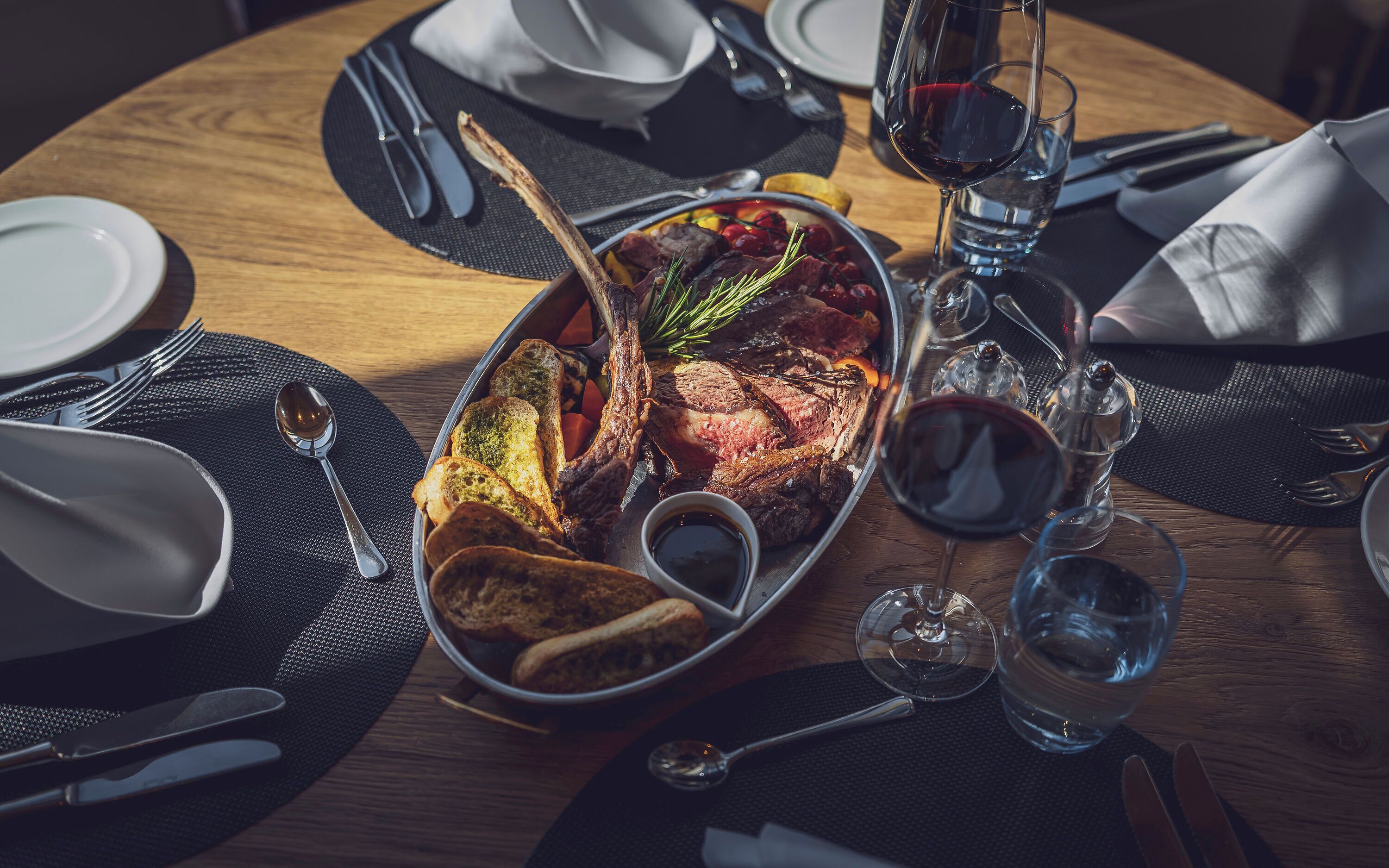 Une table à manger dressée avec des assiettes, des couverts, un verre de vin rouge et un plat de viande rôtie accompagnée de légumes.