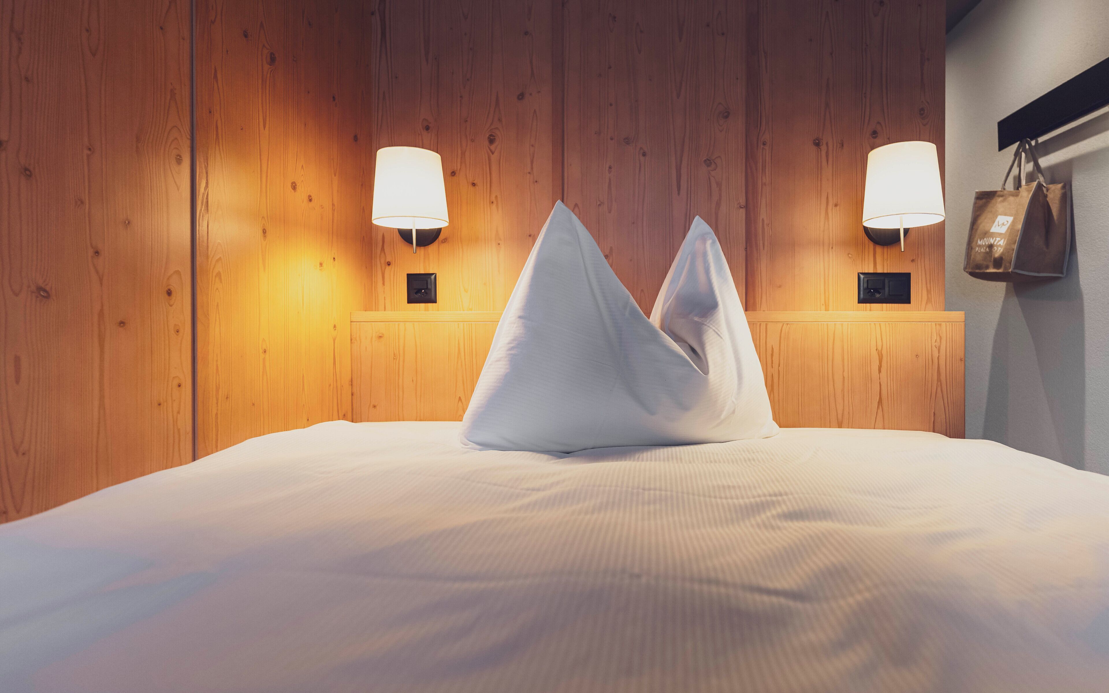 A hotel room with wooden panel walls, a bed with white linens, two wall-mounted lamps, and a bag hanging on the wall.