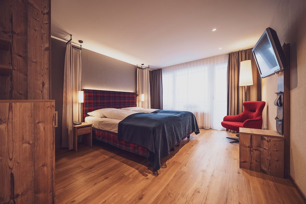 Ein Hotelzimmer mit Holzboden, einem Bett mit kariertem Kopfteil, einem roten Sessel, einem an der Wand befestigten Fernseher und einem Nachttisch mit Lampe.