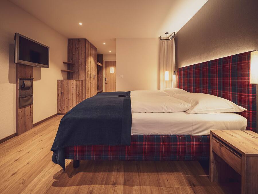 Una habitación de hotel con una cama, una mesita de noche de madera, un televisor montado en la pared y un cabecero estampado con cuadros.
