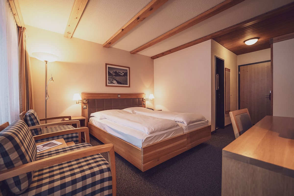 Ein Hotelzimmer mit einem hölzernen Bett, einem Schreibtisch aus Holz, einem Stuhl, einer Lampe und einem Gemälde an der Wand.