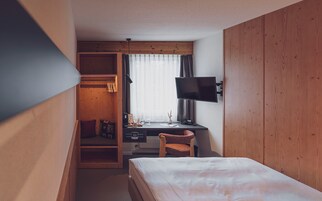Quarto de hotel com cama, escrivaninha com televisão, cadeira, guarda-roupa e janela com cortinas.