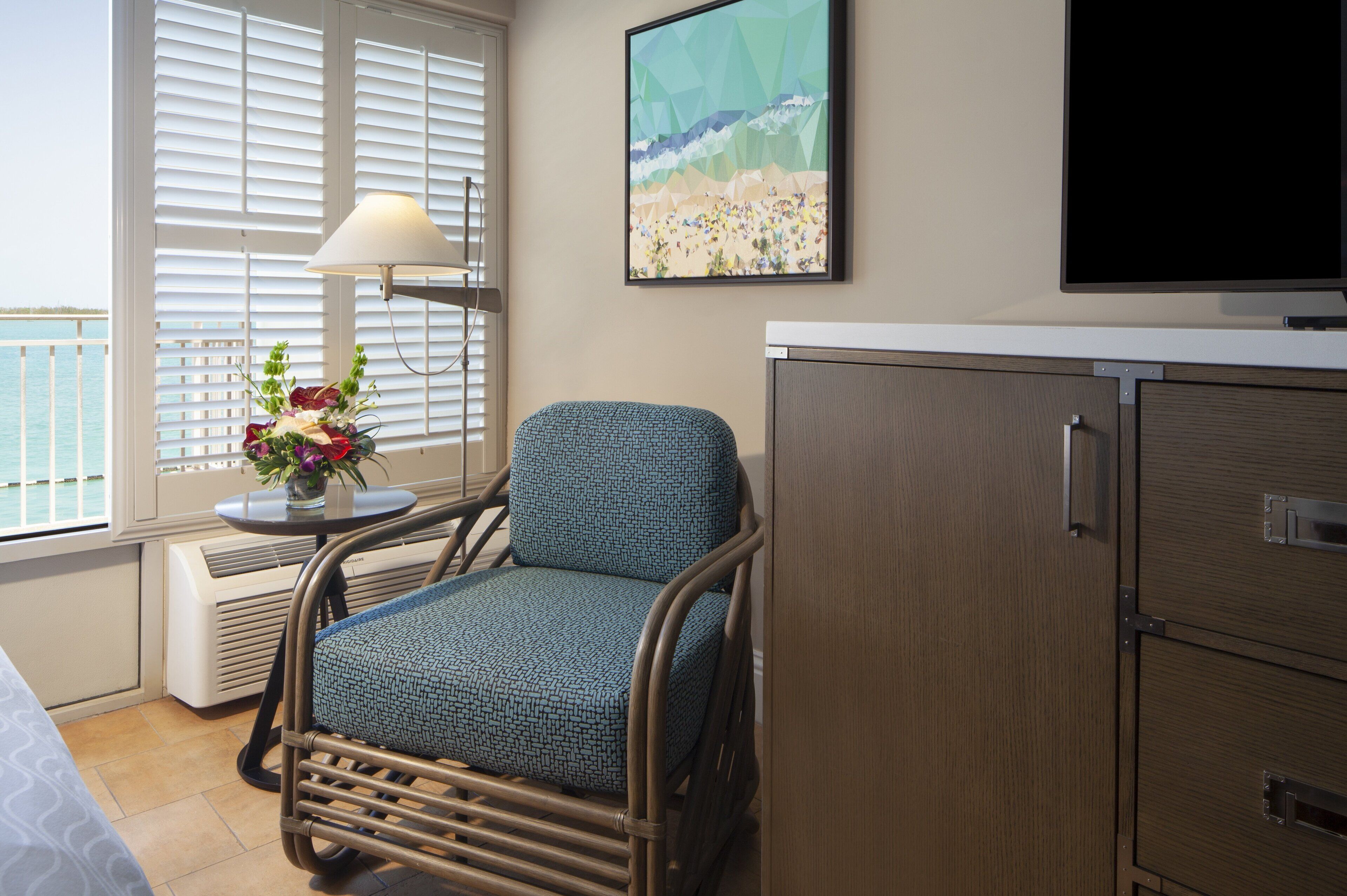 A room with a view of the sea, a chair with a patterned cushion, a bedside table with a lamp and flowers, and a cabinet.