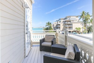 A balcony with wicker furniture, a view of the ocean, and a building with balconies and palm trees.