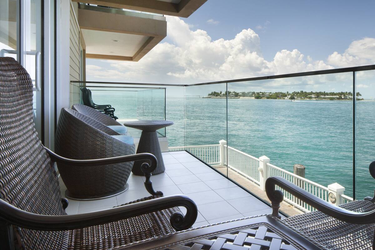 A balcony with wicker furniture, a glass railing, and a view of the sea and distant island.