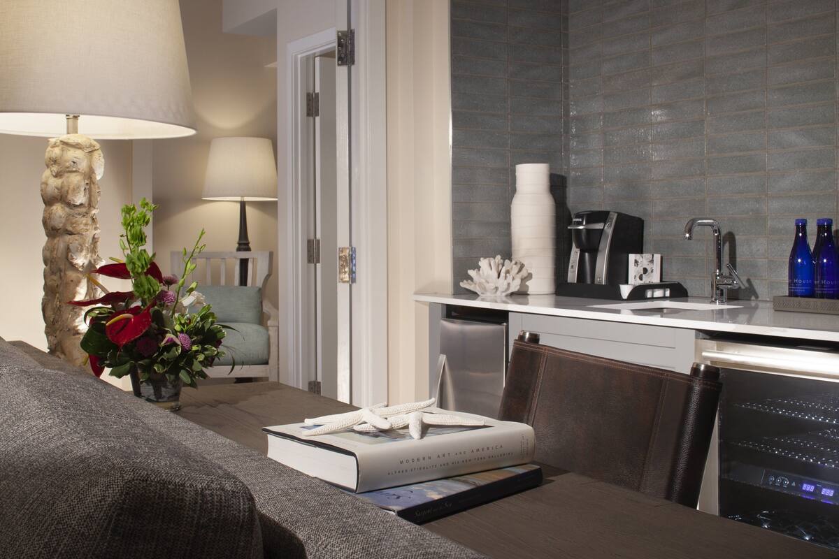 A modern hotel room with a sofa, a coffee machine, a sink, and a dining area.