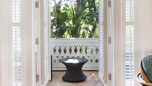 Vista através de uma janela emoldurada por branco, com uma cerca branca decorativa, que dá para um pátio pavimentado com uma mesa redonda.