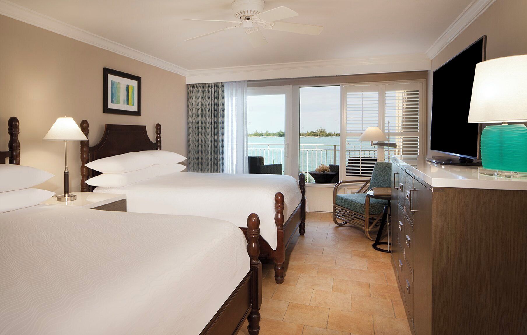 A hotel room with two beds, a television, a desk, and a view of the sea.