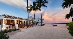 A beachfront hotel with a covered patio, palm trees, and a boat on the sand.