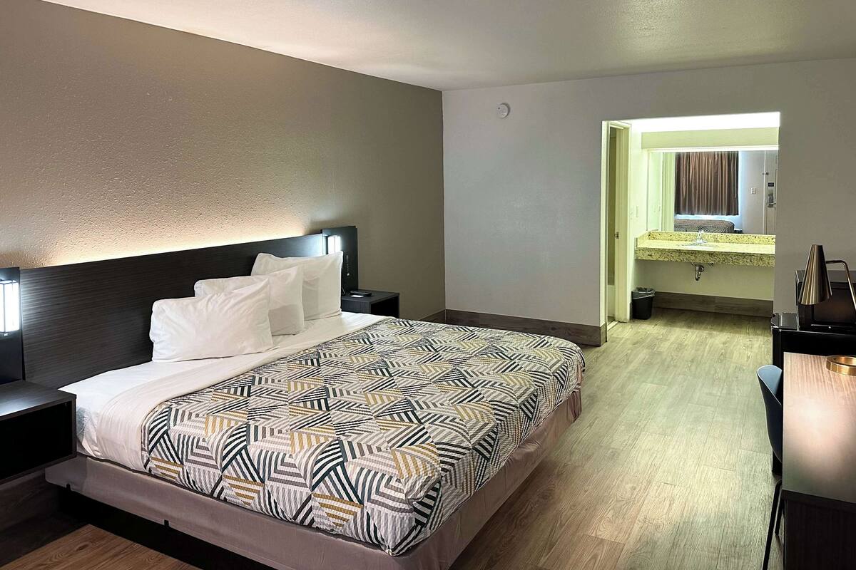 A hotel room with a large bed, a bedside table, a nightstand, a desk, a chair, a mirror, and a bathroom with a sink and toilet.