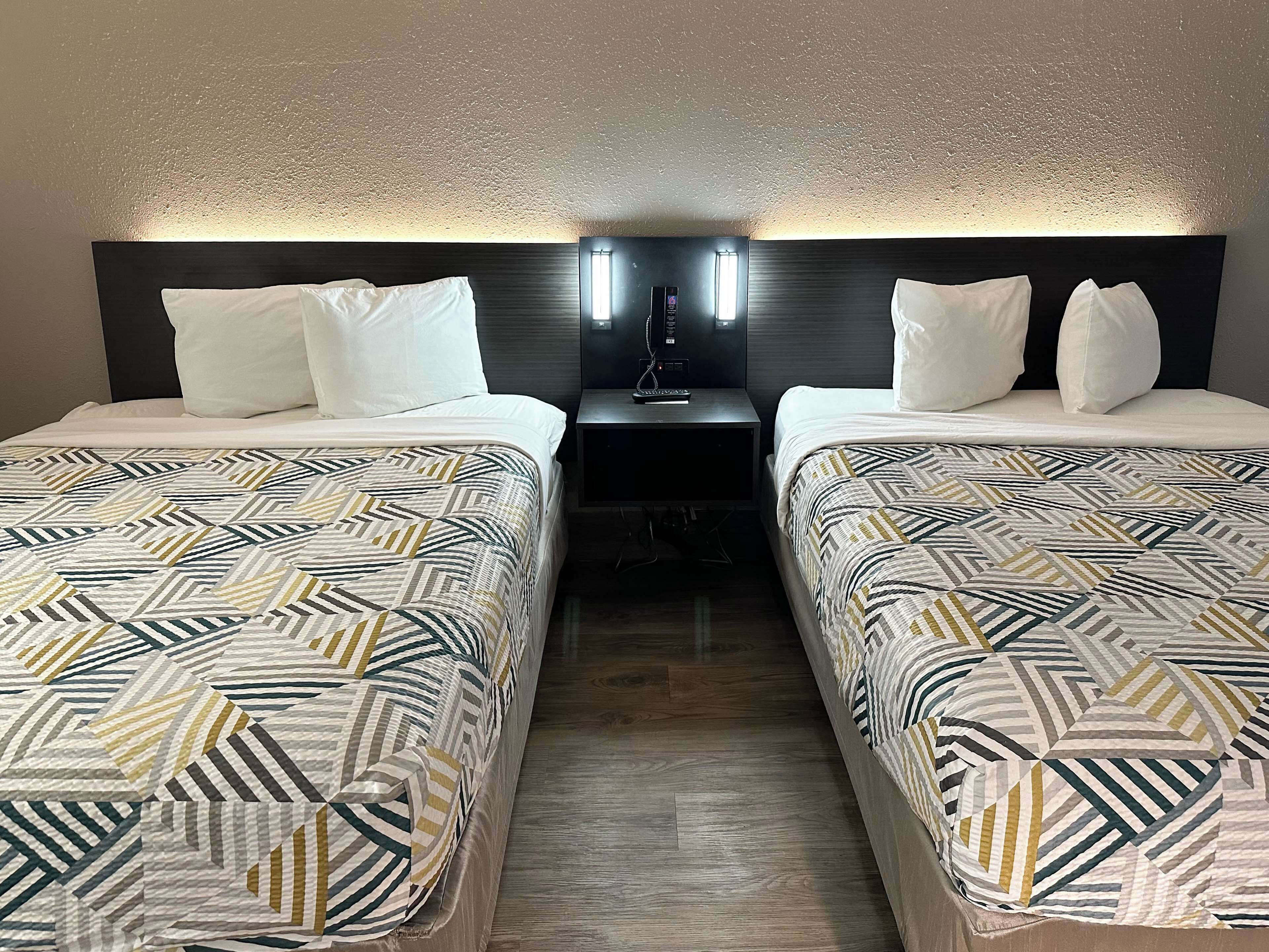 Habitación de hotel con dos camas, cada una con ropa de cama estampada, una mesita de noche con lámpara y un aplique en la pared.