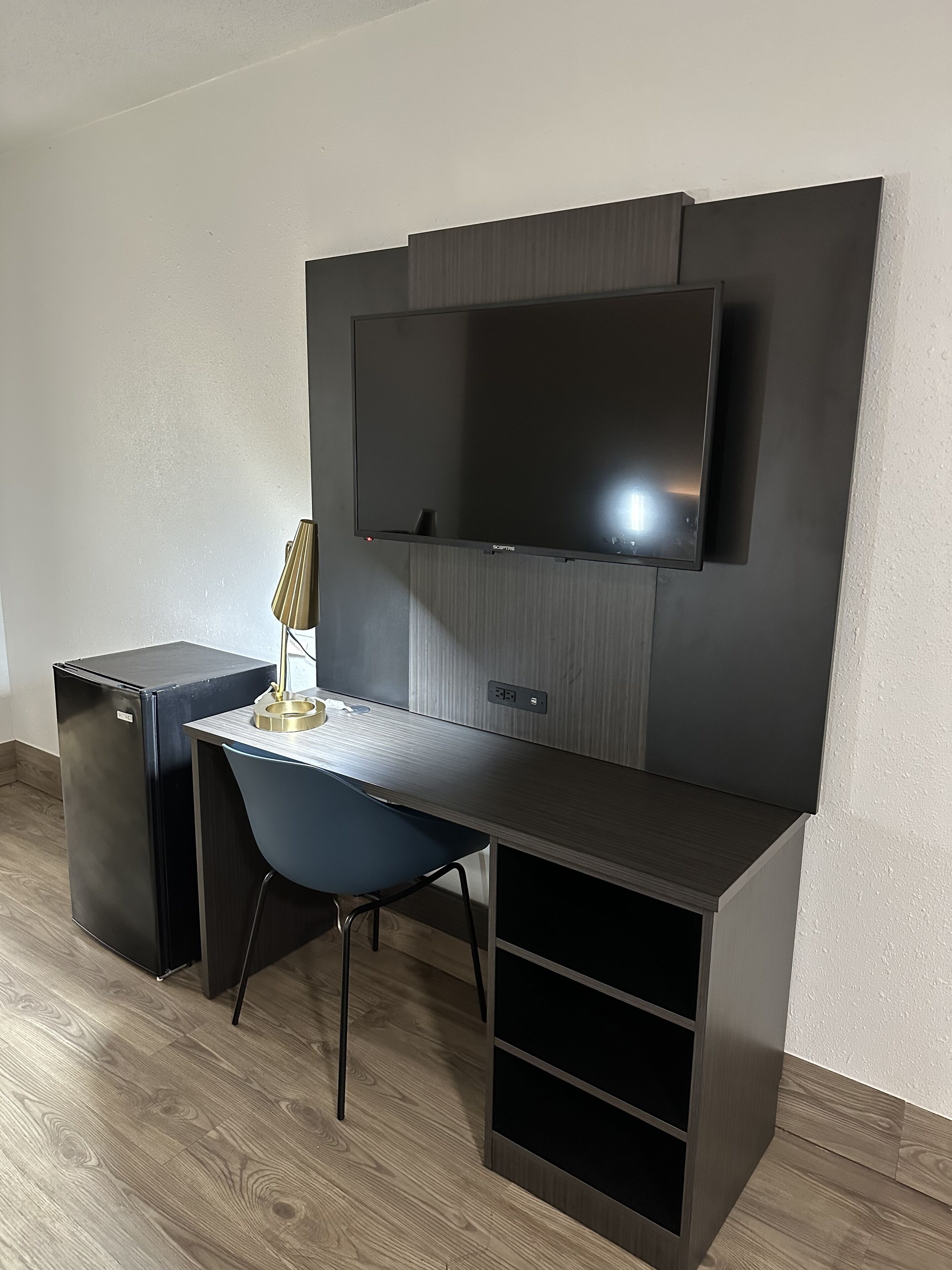 Un espace de travail moderne et compact, doté d’un téléviseur à écran plat fixé au mur, d’un petit bureau avec une chaise et d’un réfrigérateur de petite taille.