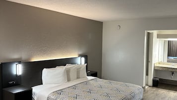 A modern hotel room with a large bed, a nightstand, a wall-mounted lamp, and a wooden floor.
