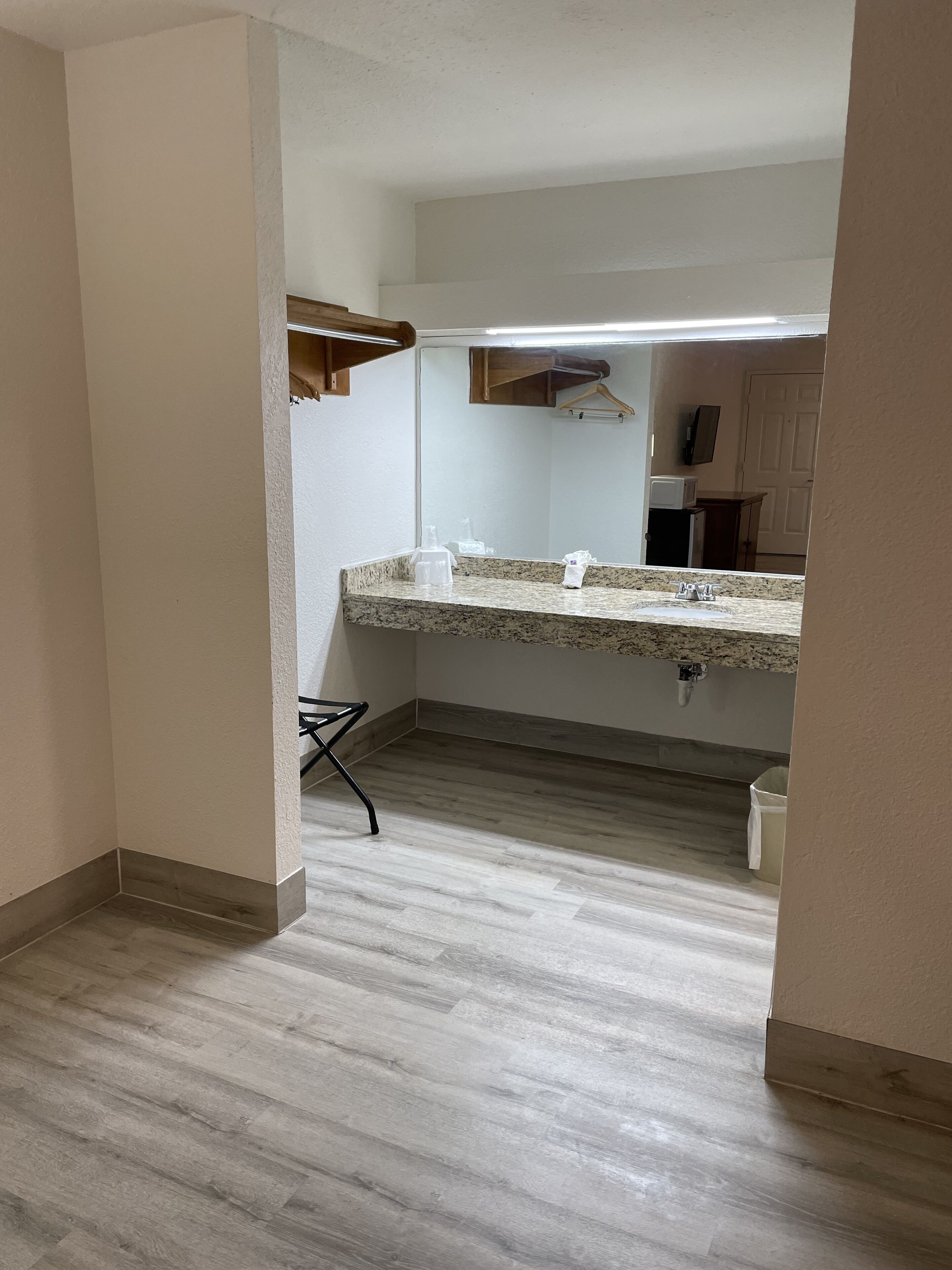 A small kitchen with a granite countertop, a sink, and a mirror above it. There is a wooden cabinet below the countertop and a small trash bin to the right.