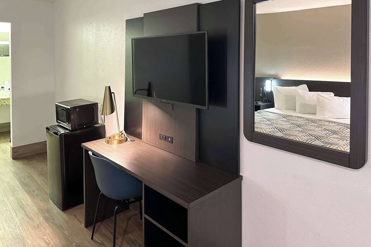 A compact hotel room with a desk, a flat-screen TV, a compact kitchenette, and a bed with a patterned headboard.