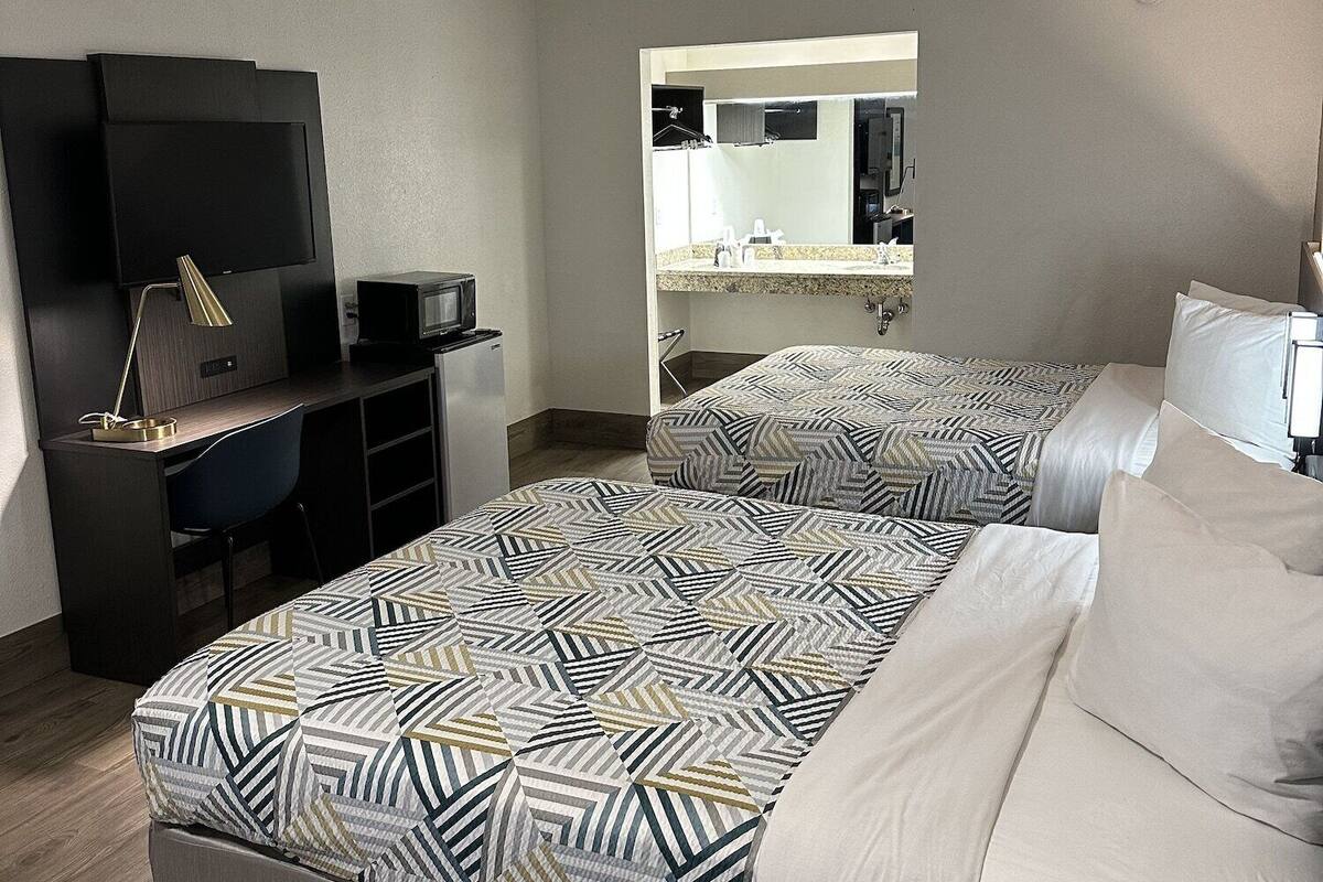 A hotel room with two beds, a desk with a computer, a TV, and a bathroom visible through an open door.