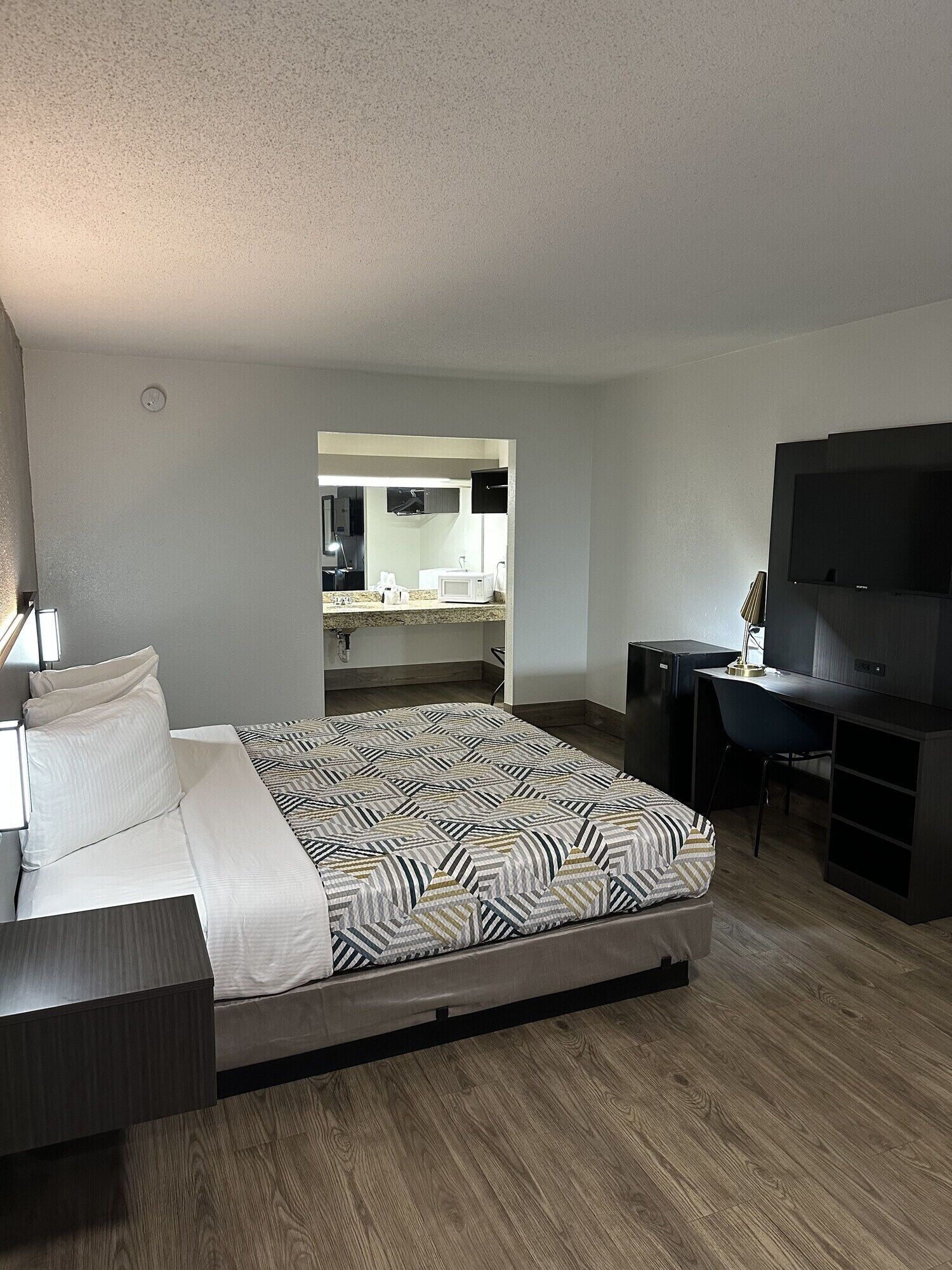 Habitación de hotel con cama, escritorio, televisor y una kitchenette al fondo.