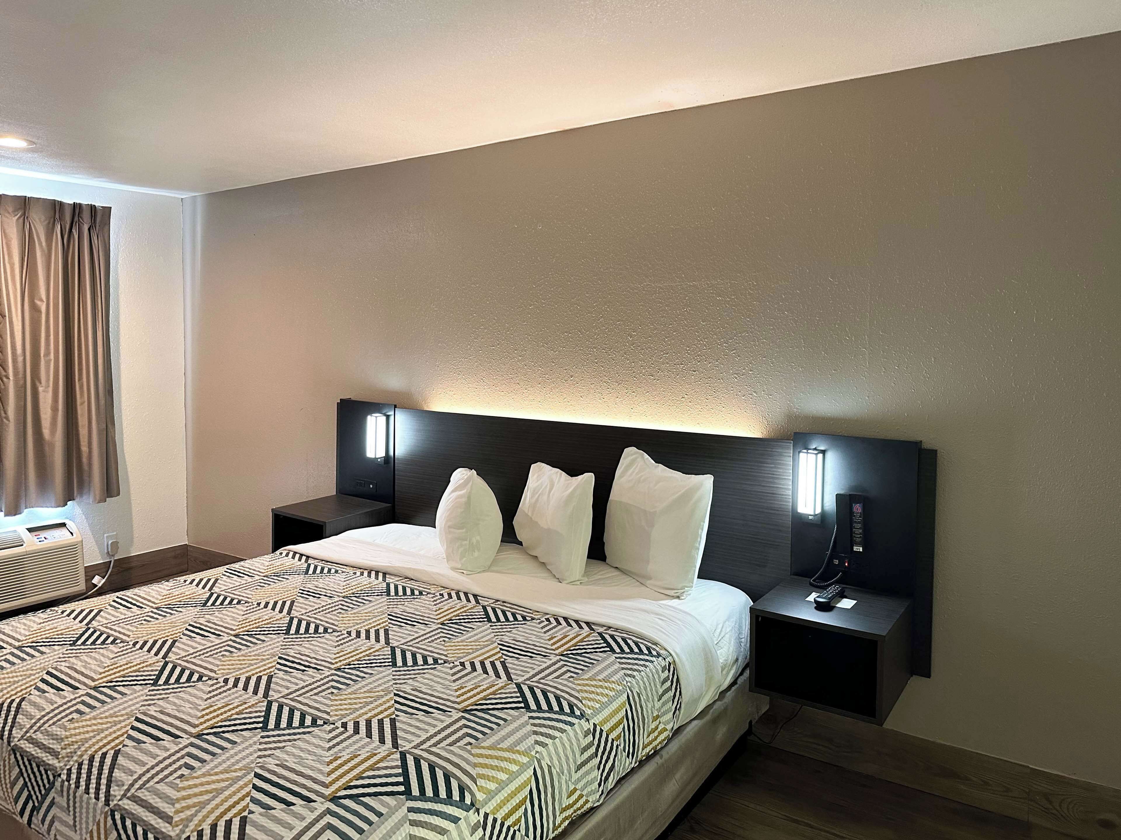 A hotel room with a large bed, two bedside tables, a headboard with integrated lighting, a window with curtains, and a wall-mounted telephone.
